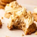 A Carrot Cake Cookie with chopped nuts sits on parchment paper, its frosted top enticingly bitten. Other similar cookies are visible in the background.