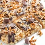 Chocolate Peanut Butter Rice Krispies treats topped with mini peanut butter cups, pretzel pieces, and drizzled chocolate are arranged on a white surface.