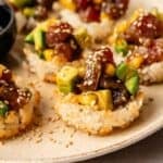 Spicy Tuna Crispy Rice rounds topped with diced avocado, tuna, mango, and sesame seeds are artfully arranged on a beige plate.