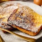 Two slices of classic French Toast dusted with powdered sugar rest on a white plate, accompanied by a gold fork and knife.