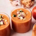 Three glasses of gazpacho topped with diced cucumber and red onion, surrounded by fresh tomatoes, bread, and garlic in the background.