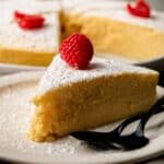 A slice of powdered sugar-dusted Japanese Cheesecake topped with a raspberry is served on a plate with two black spoons; the whole cake is in the background.
