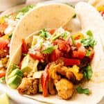 Two soft tacos filled with seasoned chicken, diced avocado, tomatoes, cilantro, bell peppers, and crumbled cheese—just like classic Sheet Pan Fajitas—served on a white plate.