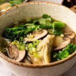 A bowl of Wonton Soup with dumplings, sliced mushrooms, broccolini, fresh cilantro, and chopped green onions in a clear broth.