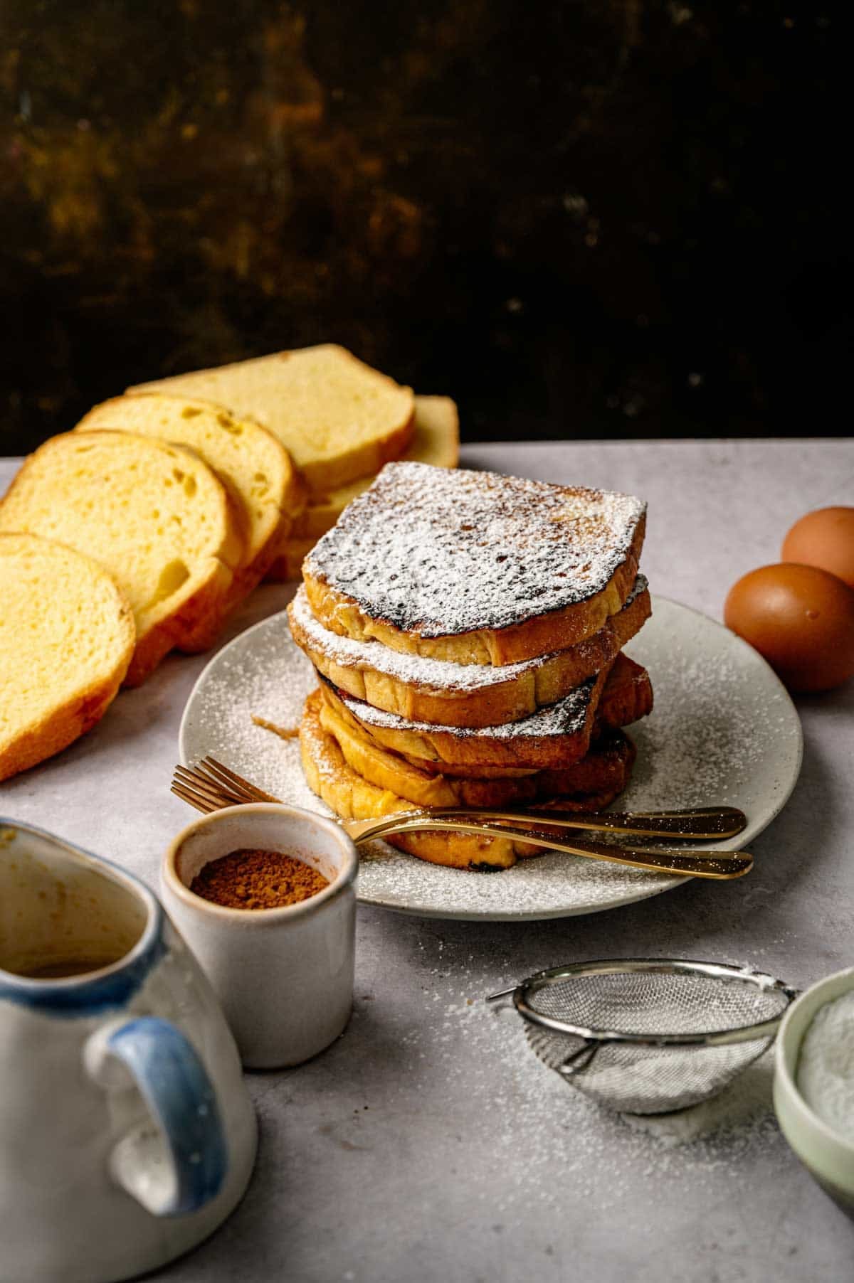 A stack of French Toast dusted with powdered sugar sits on a plate with forks, surrounded by sliced bread, eggs, a sifter, a mug, and a small container of cinnamon.