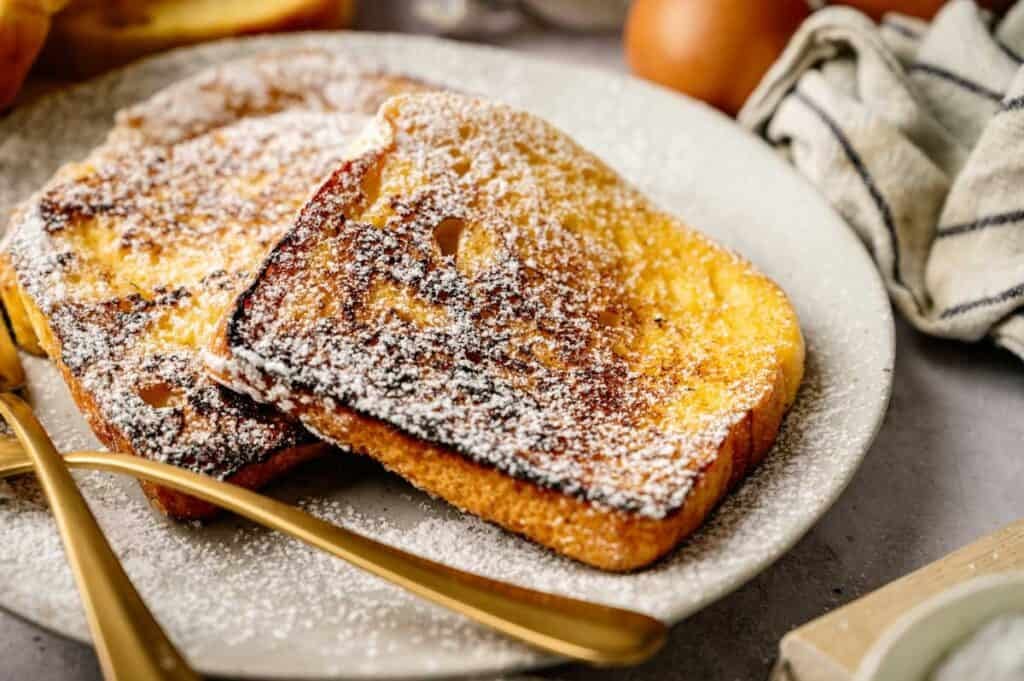 Two slices of classic French Toast topped with powdered sugar, served on a white plate with elegant gold-colored utensils.