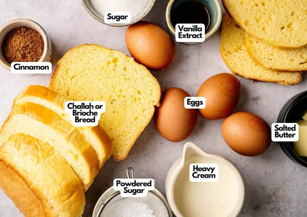 Slices of challah bread, eggs, heavy cream, salted butter, cinnamon, sugar, powdered sugar, and vanilla extract arranged on a light surface and labeled&mdash;everything you need for delicious French Toast.