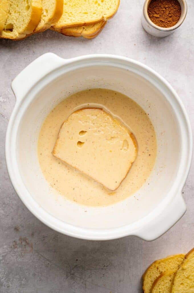 A slice of bread is soaking in a bowl of egg and milk mixture for French Toast, with a small bowl of cinnamon and more bread slices nearby on a gray surface.