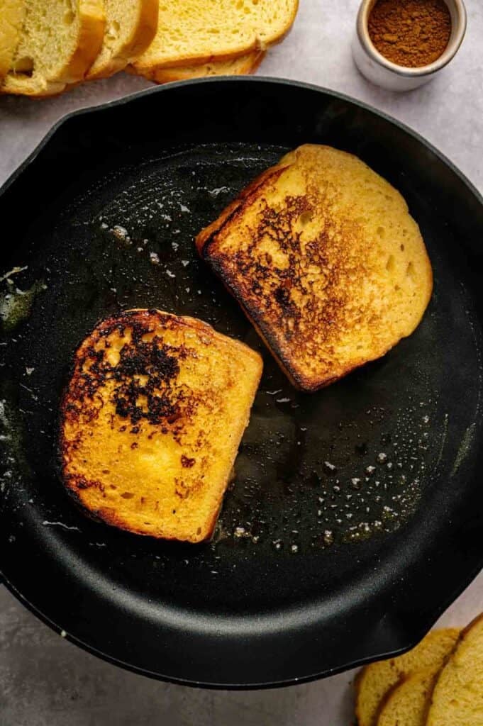 Two slices of French Toast sizzle in a black skillet, their surfaces perfectly browned; sliced bread and a small bowl of cinnamon rest nearby, ready for more delicious servings.