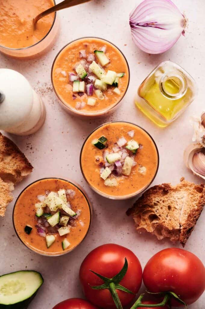 Three glasses of gazpacho topped with diced cucumber and red onion, surrounded by tomatoes, bread, oil, onion, garlic, and a pepper grinder on a light surface, showcase the fresh ingredients in this classic gazpacho recipe.