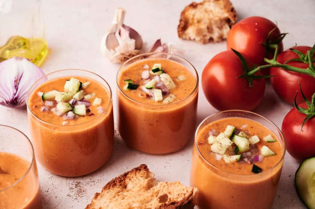 Three glasses of gazpacho topped with diced cucumber and red onion, surrounded by fresh tomatoes, garlic, bread, and olive oil on a light surface—perfect for a refreshing gazpacho appetizer.