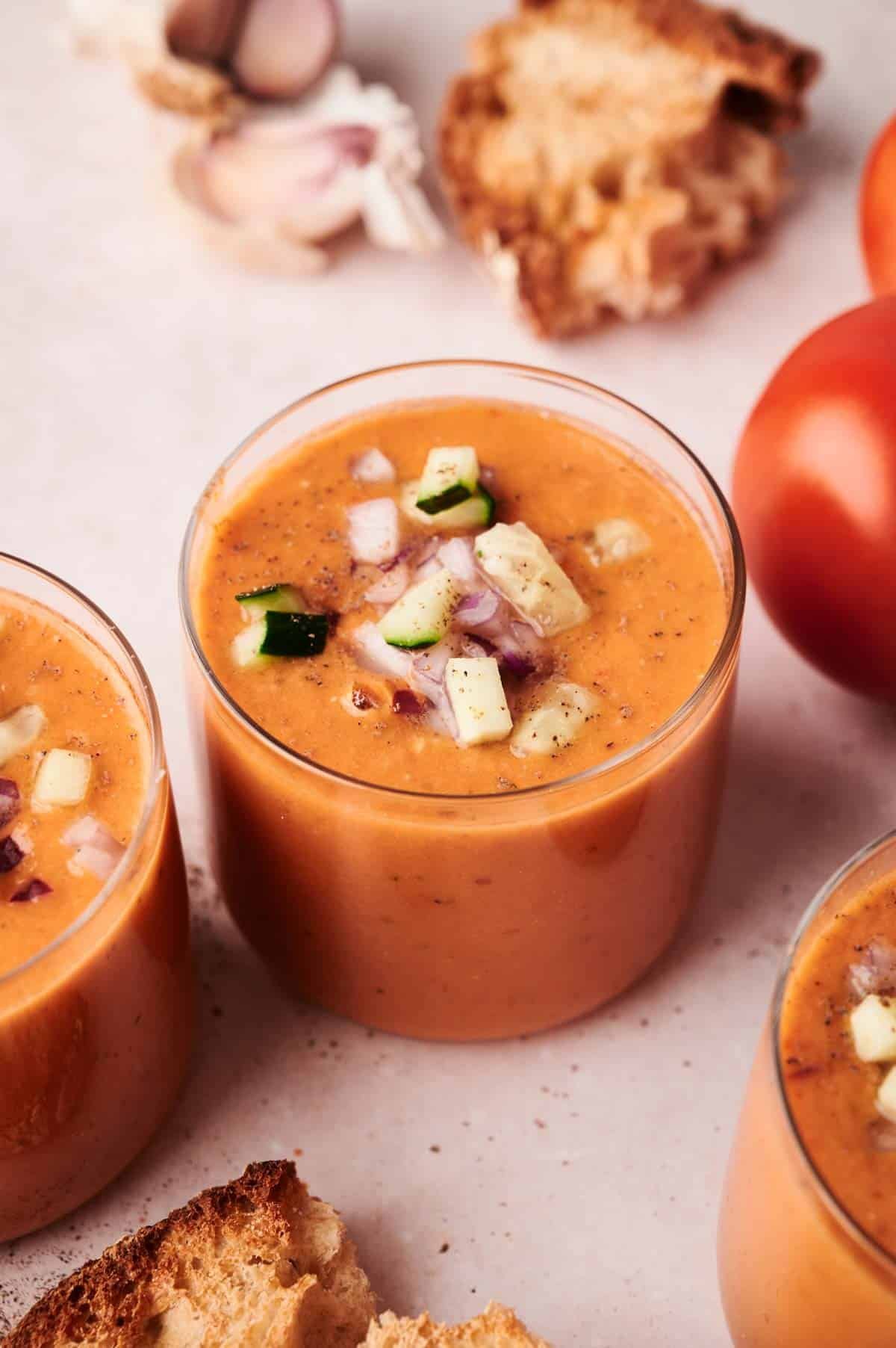 A glass of gazpacho topped with diced cucumber and onion sits surrounded by bread, garlic cloves, and whole tomatoes on a light surface. This classic gazpacho presentation highlights fresh, vibrant ingredients.
