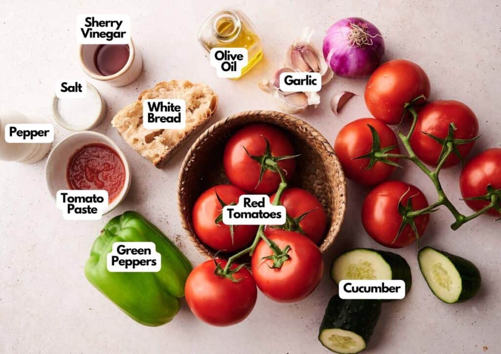 A colorful array of gazpacho ingredients—red tomatoes, cucumber, green pepper, white bread, tomato paste, salt, pepper, sherry vinegar, olive oil, and garlic—are artfully arranged on the counter.