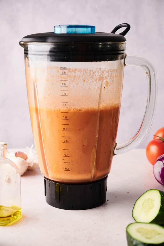A blender filled with a thick, orange-brown gazpacho sits on a countertop, surrounded by a cucumber, olive oil, tomatoes, garlic, and a red onion.
