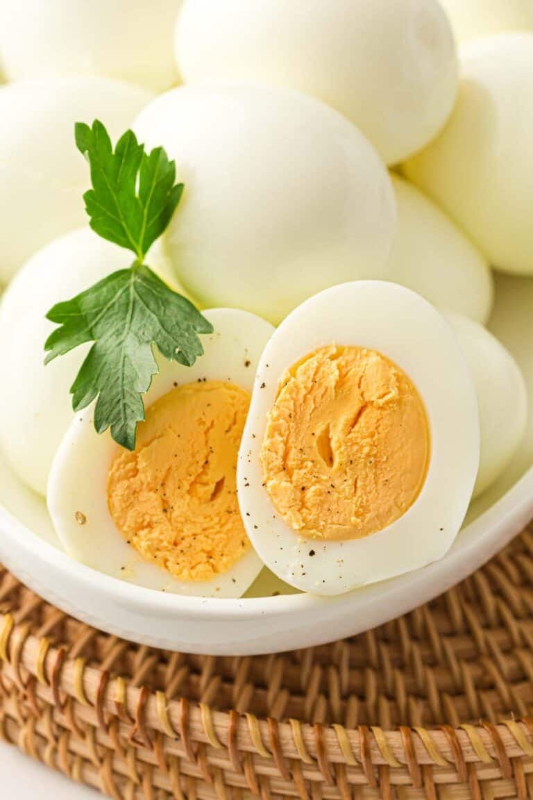 A bowl of Instant Pot Hard-Boiled Eggs, peeled and perfectly cooked, with one egg cut in half, garnished with a sprig of parsley and a sprinkle of black pepper.