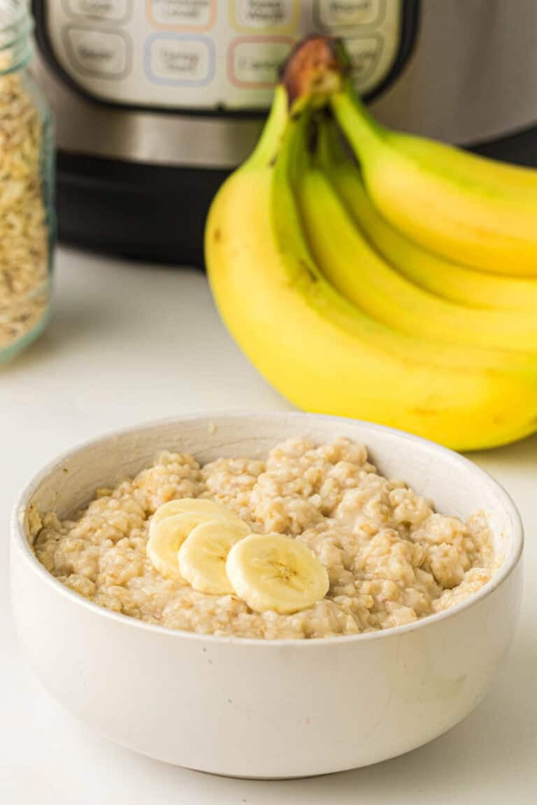 A bowl of Instant Pot Oatmeal topped with banana slices sits in front of a bunch of bananas and an electric pressure cooker.
