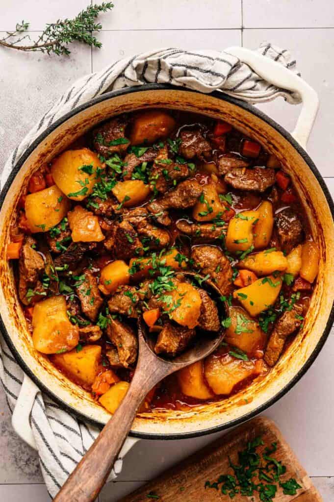 A pot of Irish Stew with chunks of beef, potatoes, carrots, and garnished with chopped parsley, with a wooden spoon resting inside.