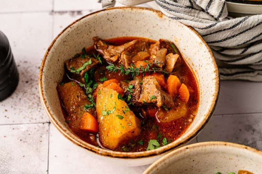 A bowl of Irish Stew with chunks of beef, potatoes, carrots, and herbs in a rich broth, garnished with fresh parsley and thyme.