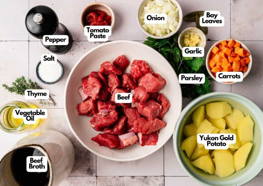 A top-down view of ingredients for Irish Stew, including cubed beef, potato, carrot, onion, garlic, parsley, thyme, bay leaves, tomato paste, salt, pepper, vegetable oil, and beef broth.