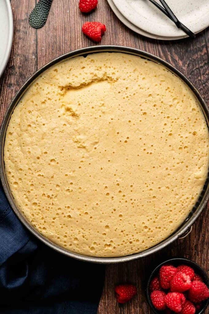A round, golden Japanese Cheesecake in a baking pan sits on a wooden table next to a small bowl of fresh raspberries and a white plate.