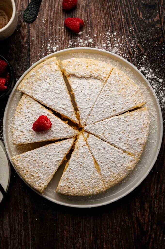 A round Japanese Cheesecake dusted with powdered sugar, sliced into eight pieces, with one slice topped by a fresh raspberry, on a white plate on a wooden table.