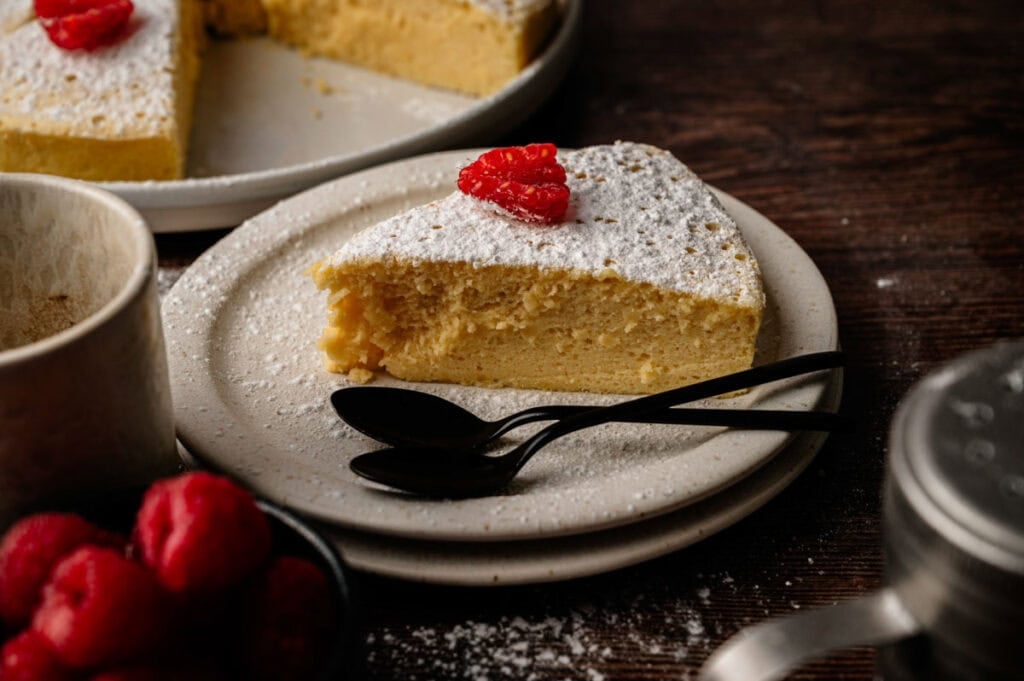 A slice of Japanese Cheesecake topped with powdered sugar and raspberries rests on a plate with two black spoons, with more cheesecake and fresh raspberries in the background.