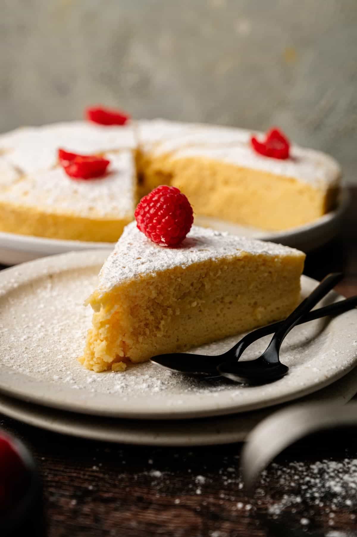 A slice of Japanese cheesecake topped with powdered sugar and a raspberry sits on a plate with two black forks; more cake is in the background.