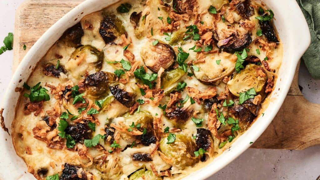Oval baking dish filled with roasted Brussels sprouts casserole, topped with melted cheese, crispy onions, and chopped parsley.