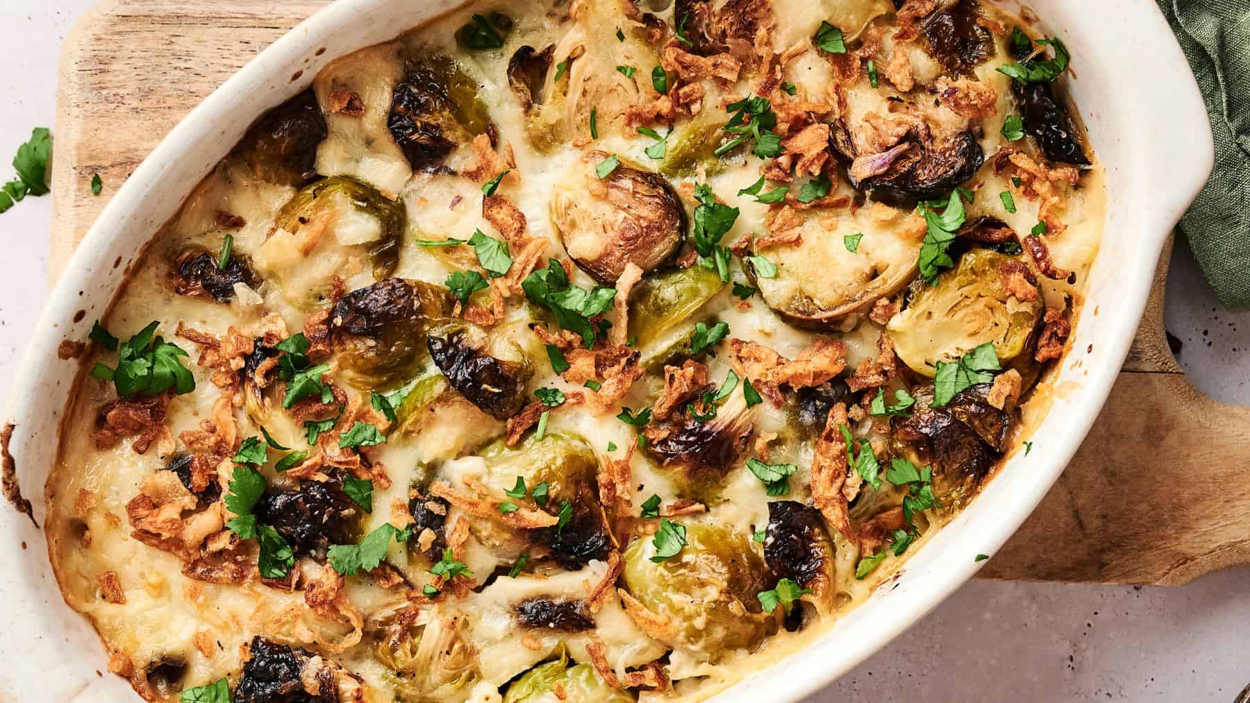 A baked casserole dish filled with roasted Brussels sprouts, melted cheese, crispy onions, and chopped herbs on top.