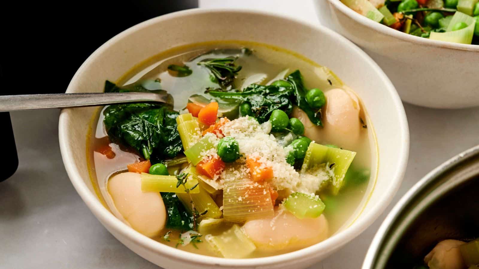 A bowl of vegetable soup with spinach, peas, carrots, celery, and large white beans, topped with grated cheese.