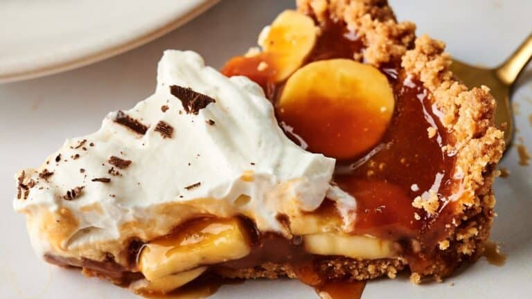 A slice of banoffee pie with a crumbly crust, banana slices, caramel, chocolate shavings, and a topping of whipped cream on a white plate.