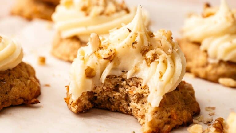 A close-up of a frosted cookie topped with chopped nuts, with a bite taken out of it. Other similar cookies are visible in the background.