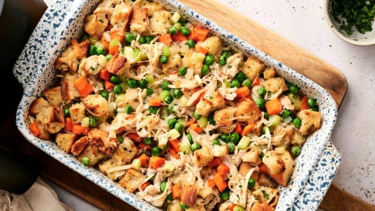 A casserole dish filled with a baked mixture of bread cubes, shredded chicken, carrots, celery, and green peas on a wooden board.