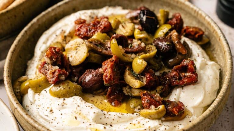 A bowl of whipped feta cheese topped with chopped olives, sun-dried tomatoes, olive oil, and black pepper.