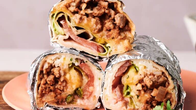 Three foil-wrapped burritos cut in half, showing ground meat, lettuce, tomato, cheese, and sauce, stacked on a pink plate.