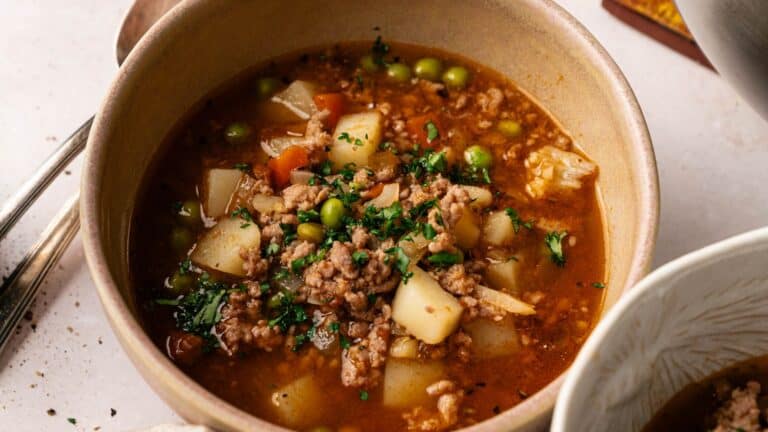 A bowl of ground beef stew with potatoes, peas, carrots, and a tomato-based broth, garnished with chopped herbs.