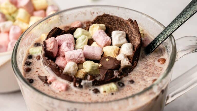 A glass mug of hot chocolate topped with a chocolate shell and colorful mini marshmallows, with a spoon resting inside.