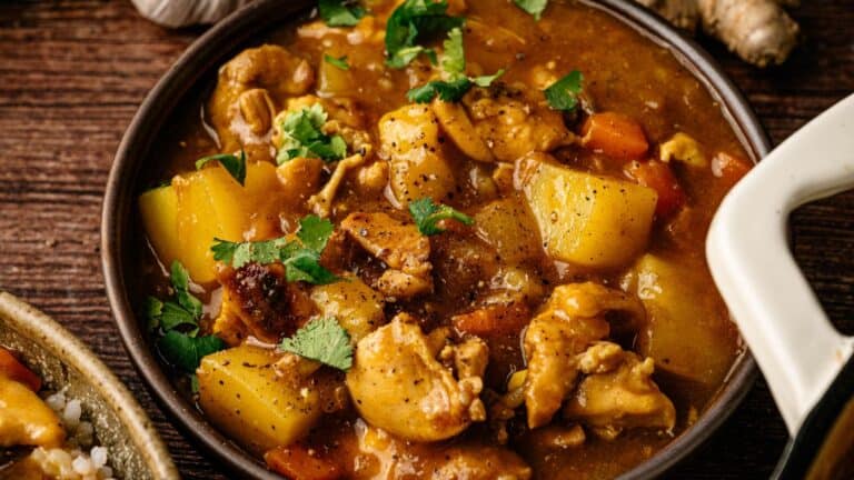 A bowl of chicken curry with potatoes, carrots, and fresh cilantro, served in a brown dish on a wooden table.