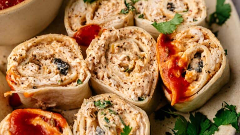 Close-up of sliced tortilla pinwheels filled with a creamy mixture, garnished with herbs and topped with a red sauce.