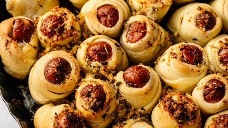 Baked pigs in a blanket with crescent roll dough wrapped around mini sausages, topped with herbs and seasoning, arranged closely together in a pan.
