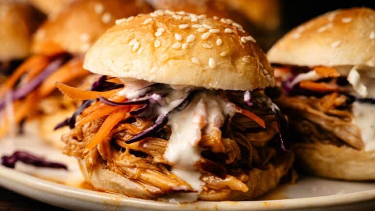 A close-up of a pulled chicken sandwich with shredded carrots, purple cabbage, and creamy sauce on a sesame seed bun.