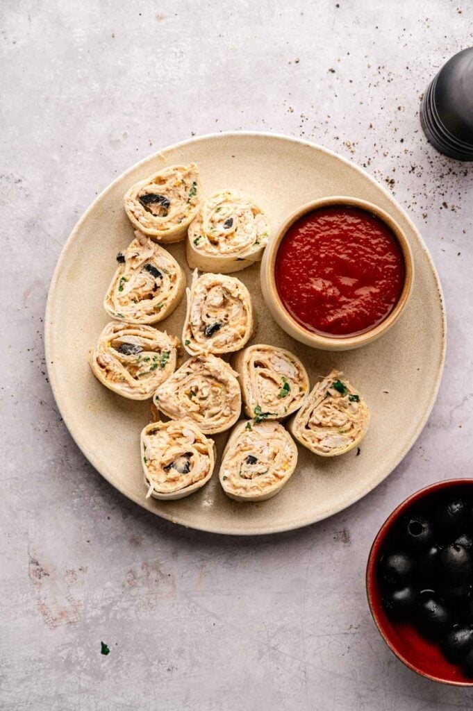 A plate of Mexican Pinwheels with a side of red dipping sauce sits next to a bowl of black olives on a light surface.