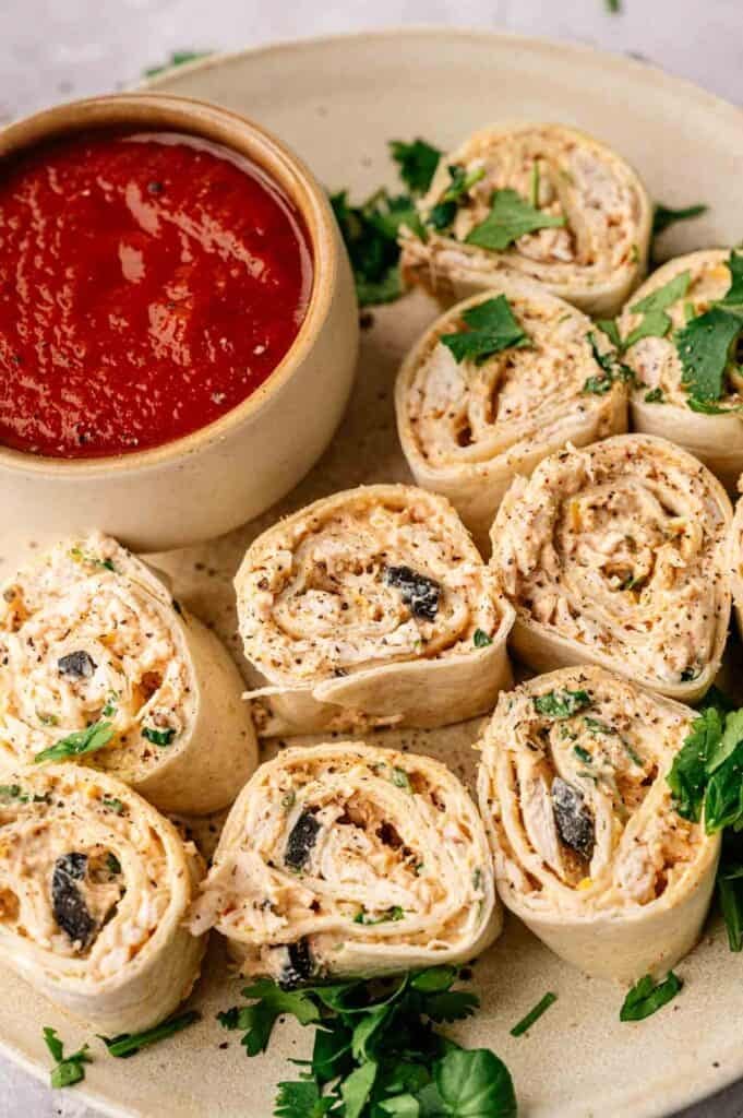 Mexican Pinwheels filled with a creamy mixture and black olives are arranged on a plate with parsley, served alongside a bowl of red dipping sauce.