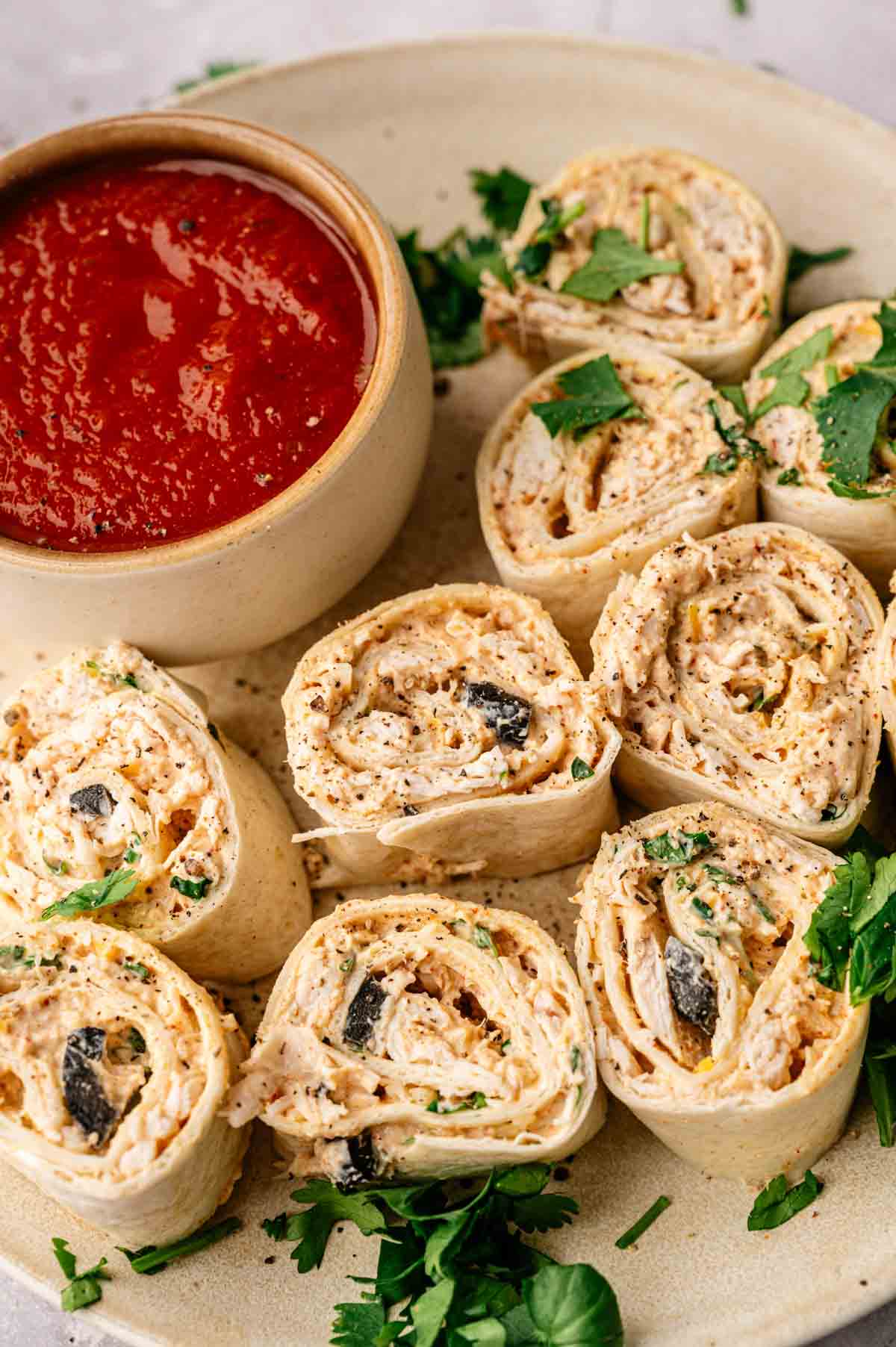 Mexican Pinwheels filled with a creamy mixture and black olives are arranged on a plate with parsley, served alongside a bowl of red dipping sauce.