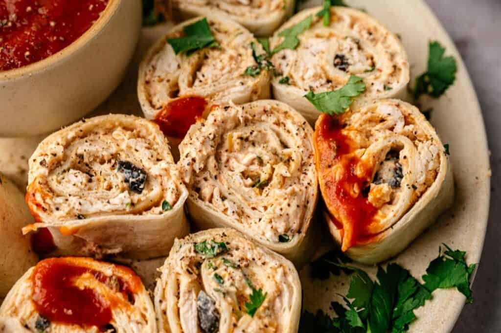 A plate of Mexican Pinwheels filled with a creamy mixture, garnished with cilantro, and served alongside a bowl of red salsa.