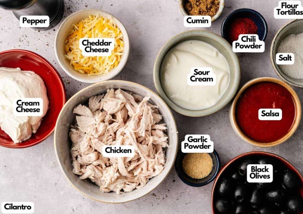 An overhead view of labeled ingredients for Mexican Pinwheels, including shredded chicken, cheeses, spices, sour cream, salsa, black olives, and flour tortillas arranged on a counter.