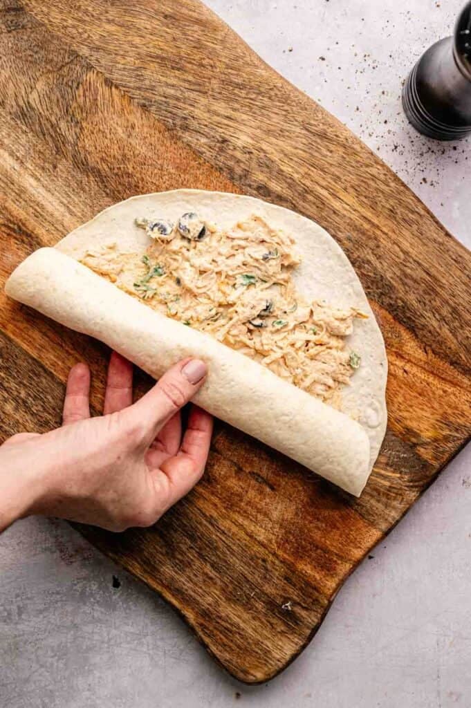 A hand rolls a flour tortilla filled with a creamy chicken mixture on a wooden cutting board, preparing delicious Mexican Pinwheels.
