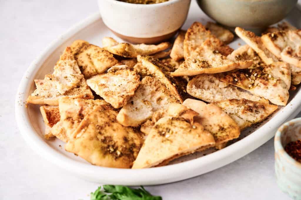 A white plate filled with crispy pita chips seasoned with spices, next to small bowls of dip.