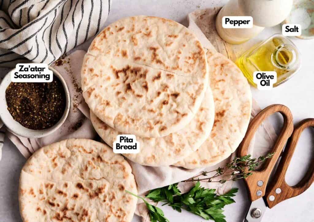 Four pieces of pita bread on a cloth, surrounded by za&rsquo;atar seasoning, olive oil, salt, pepper, kitchen scissors, and fresh herbs&mdash;perfect for making homemade pita chips.