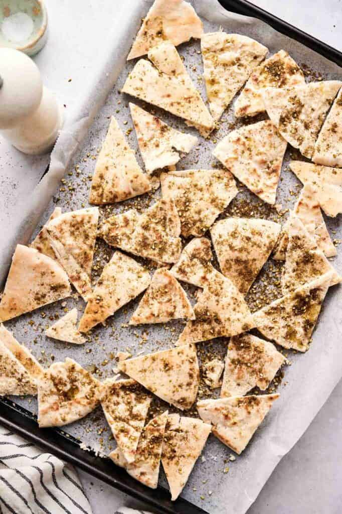 A baking sheet lined with parchment paper holds pita chips sprinkled with za'atar seasoning, ready for baking.